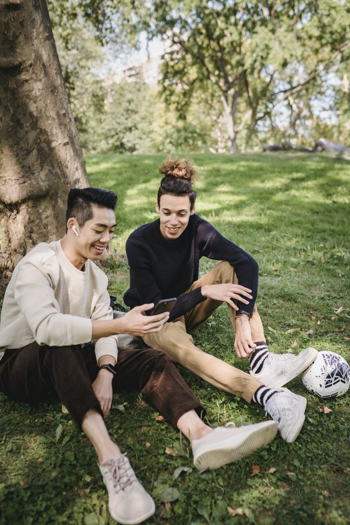 Smiling young ethnic males in casual clothes watching funny video on modern mobile phone while resting together on grass meadow in park